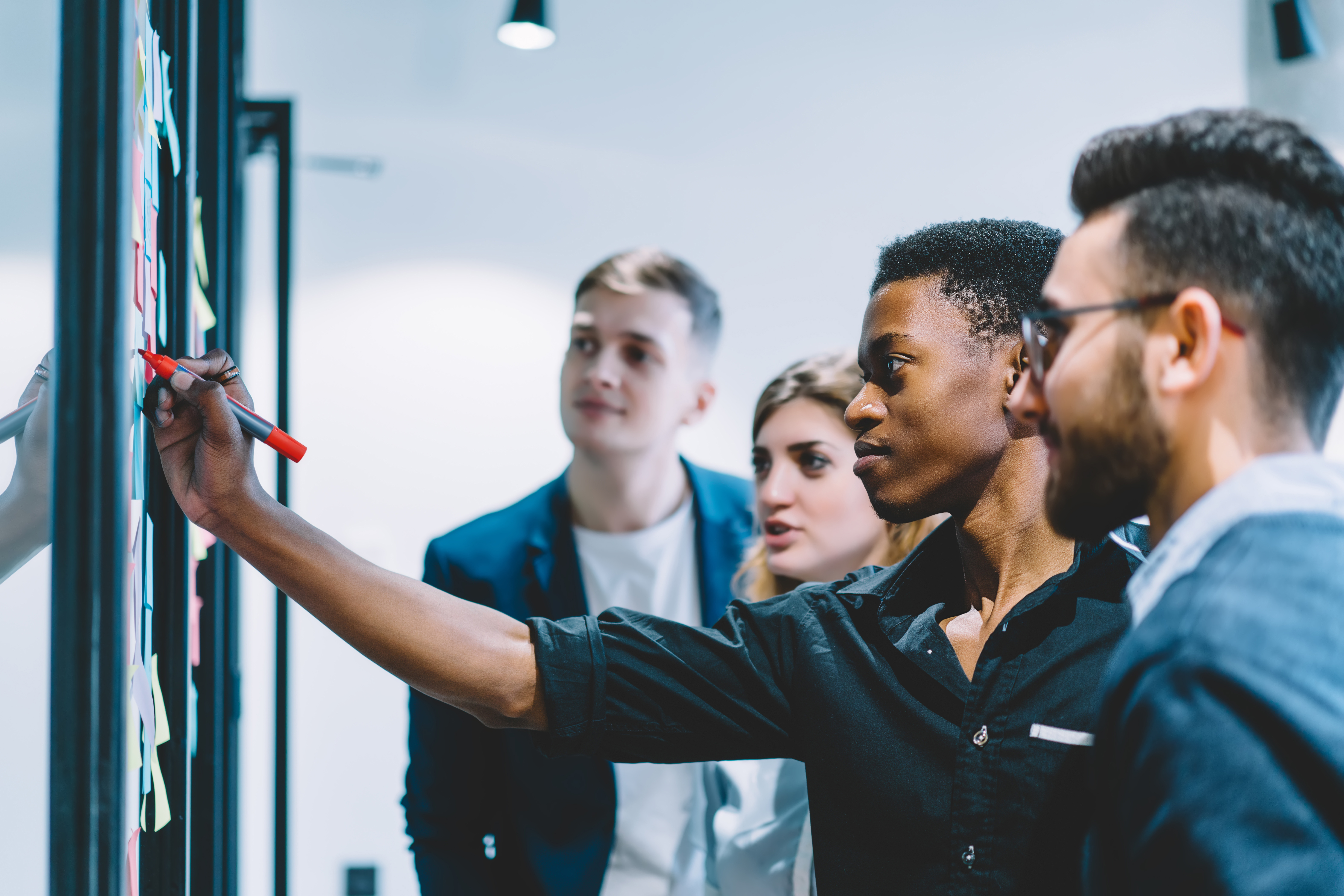 Diverse team of skilled students together solving task writing down information on papers glued on wall.Multicultural young people collaborating on business plan making notes of ideas on sticker